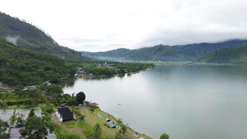 Serene lake surrounded by lush green mountains and a small village along its shoreline reflects the calm atmosphere of the cloudy day with a boat visible on the water.