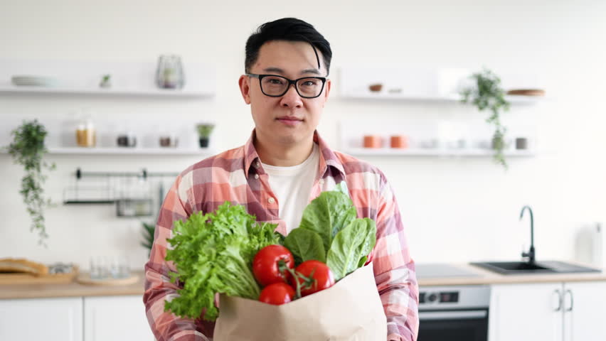 An Asian man smiles while holding a bag of fresh produce in his kitchen