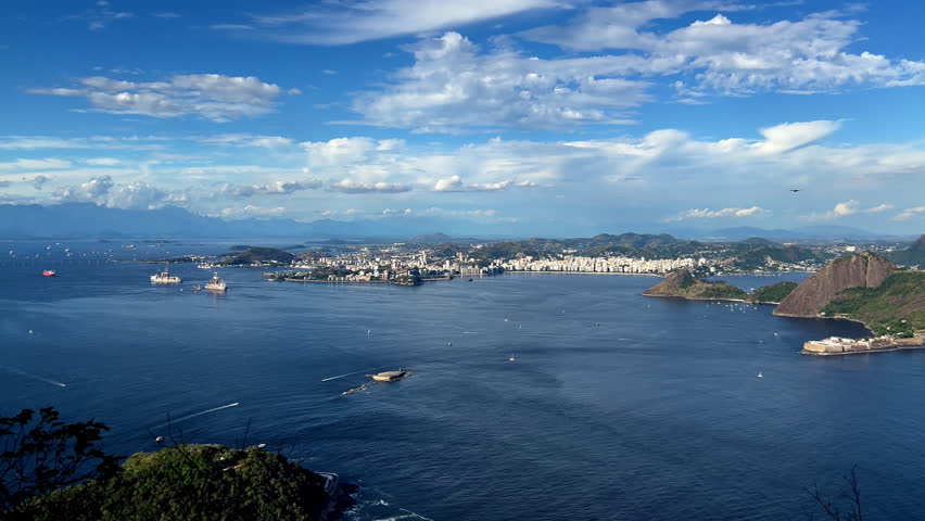 Panorama from the top of Pão de Açúcar overlooking Niterói. Captures the historic Santa Cruz Fortress, scenic Niterói beaches, and the expansive blue waters of Guanabara Bay under a clear tropical sky