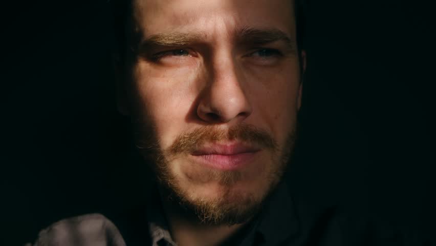 Man with beard squinting and shielding his eyes from intense sunlight, with shadows from blinds creating striped pattern across his face in dark room. Blinded man shielding eyes from bright sunlight