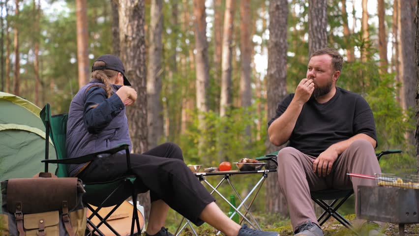 Relaxed man and woman sitting on camping chairs, eating snacks and waiting for the barbecue to cook while enjoying their vacation in a beautiful pine forest with a tent in the background