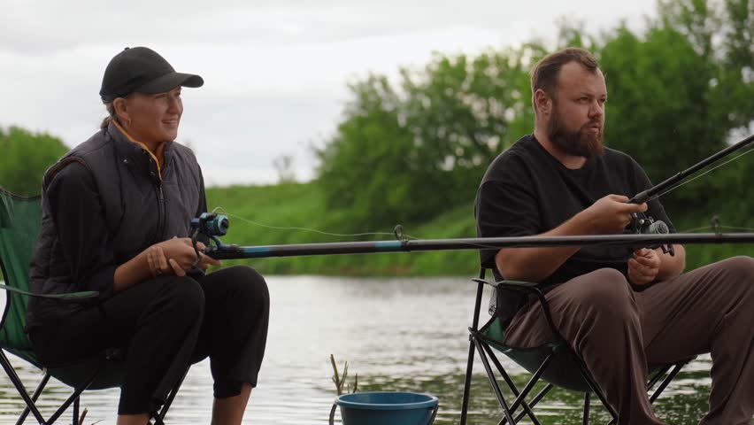 Man and woman enjoying a quiet day fishing together, sitting on folding chairs by the river with their rods in the water, relaxing during a weekend getaway in the beautiful countryside