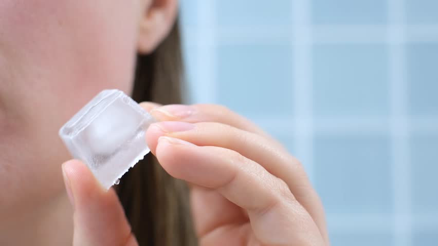 Young woman holding ice cube near her face against background of blue tiles.