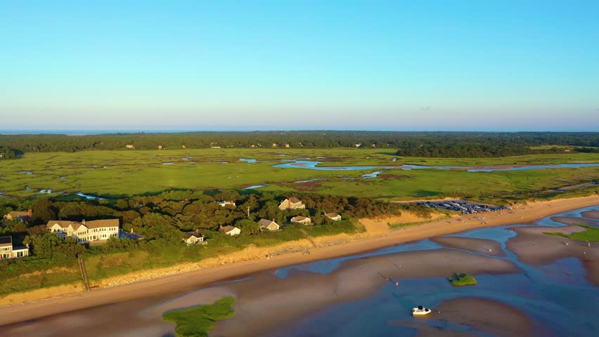 Drone sunset aerial of Cape Cod shoreline with tidal flats and shallow pools, beachgoers scattered along the sand, and green marshland. Scenic New England coast, smooth 4K b-roll.