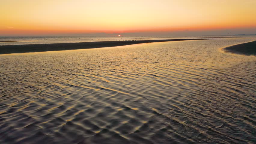 Drone aerial over low tide sandbars at sunset as shorebirds gather and glide across rippled shallows. Calm water reflects warm orange skies, creating a serene coastal seascape, cinematic 4K b-roll.