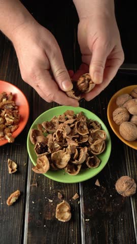 Hands of a person engaged in cracking walnut shells, with a green bowl filled with empty shells at the center, surrounded by bowls of nuts on a rustic wooden table