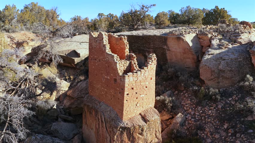 A tall rectangular stone ruin sits perched on a sandstone pedestal. The camera pulls back and tilts down to reveal the precarious masonry set against the rugged beauty of a western desert canyon.