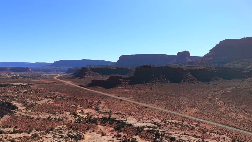 A long, straight road stretches across a vast desert valley surrounded by high red rock walls. This wide drone view captures the open space and remote beauty of a western American road trip.