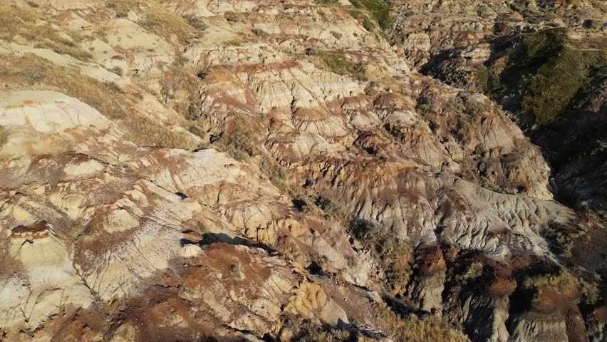 Low altitude aerial shot over eroded badlands hills in Alberta, Canada, revealing deep valleys carved between formations under strong side lighting with high contrast near sunset.