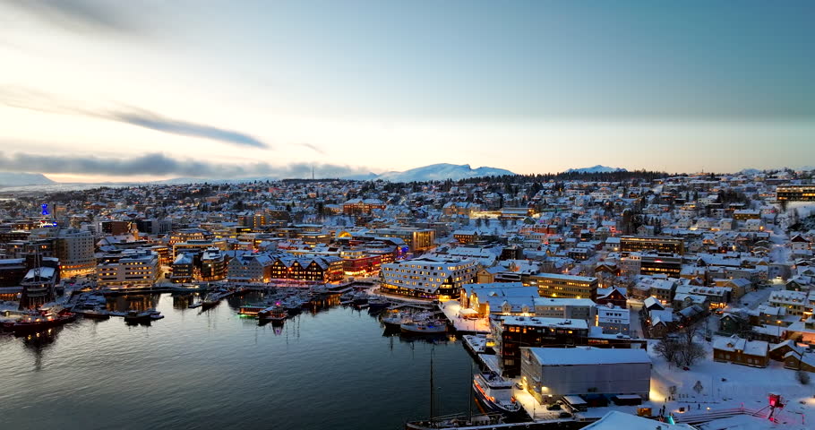 Tromsø, Northern Norway at blue hour, showing snowy rooftops, harbor water and warm city lights reflecting on the fjord in winter, aerial view
