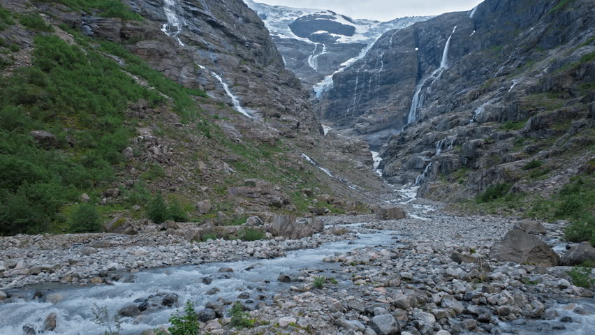 Female tourist walking toward Kjenndal Glacier in Norway. Traveler hikes across a rocky riverbed with flowing stream, waterfalls and icy glacier framed by steep mountain cliffs on a cool summer adventure.