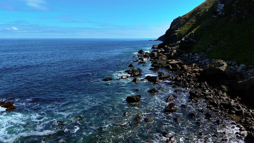 Waves rolling over black rocks along green hillside coast. Sea moving through scattered stones on rugged shore. Ocean washing against uneven rocky beach beside steep cliff