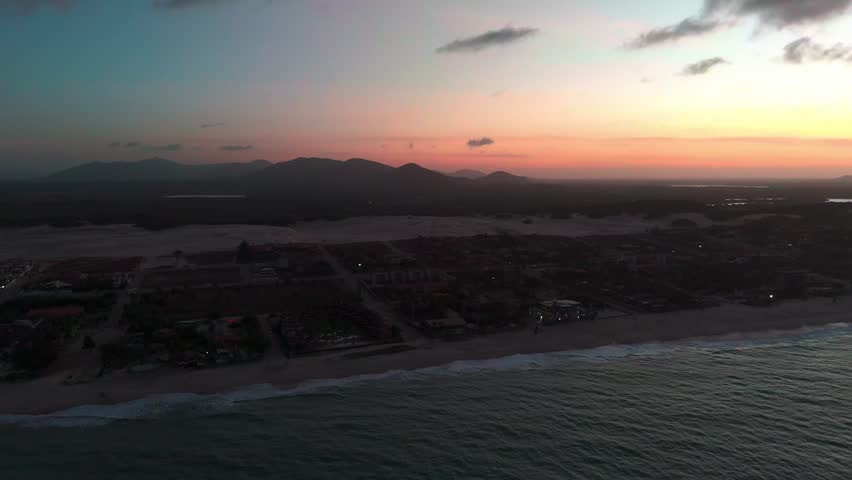 Aerial wide shot of a tropical beach with turquoise water and waves crashing on the sand, coastline resorts and sand dunes in the background, Brazil.