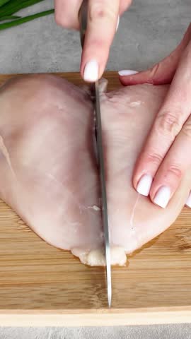 Woman cutting a fresh raw chicken breast in half with a large kitchen knife on a wooden cutting board