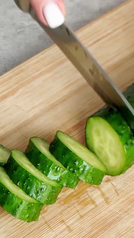 Woman's hand using a sharp knife for slicing a fresh, ripe green cucumber on a wooden cutting board. Healthy food preparation for a salad
