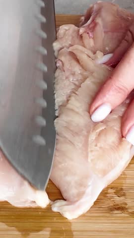 Close-up of a woman's hands cutting raw chicken breast fillet with a large kitchen knife on a wooden cutting board. Preparing poultry for cooking