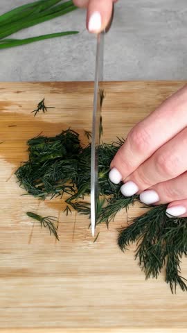Woman's hands using a large kitchen knife to chop fresh dill on a wooden cutting board. Healthy cooking and meal preparation at home