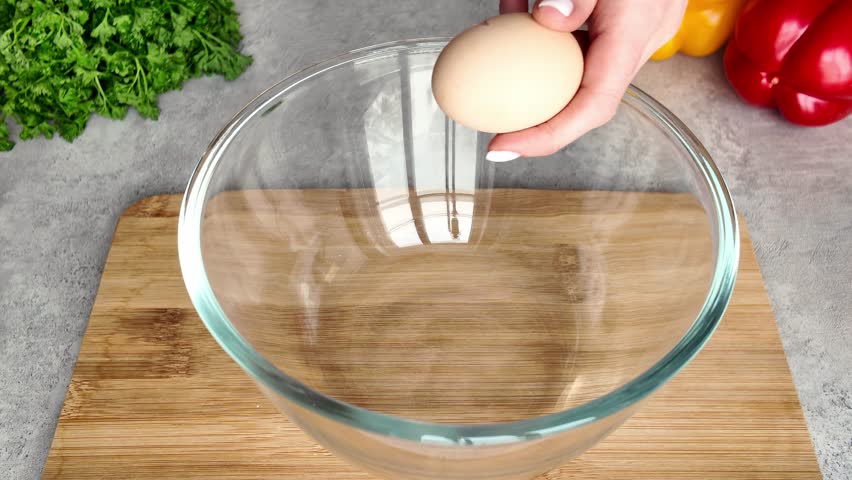 A female cook cracks a fresh raw egg with a knife over a clear glass bowl. The breakfast preparation process