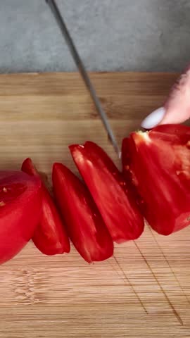 Female hands with a knife cutting a fresh red tomato into thin slices on a wooden bamboo board. Close-up of food preparation for cooking a meal