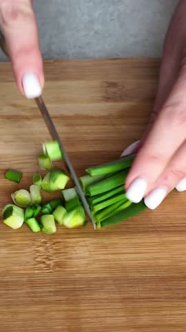 Close-up of female hands with a knife slicing fresh spring onions on a bamboo cutting board. Healthy cooking and meal preparation concept