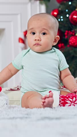 Adorable Small Bald Infant Boy In Green Bodysuit Sitting Near Festive Christmas Tree With Red Ornaments Looking Calm Sweet Serious Cute Toddler Kid Holding Colorful Gift Box Child Vertical Video