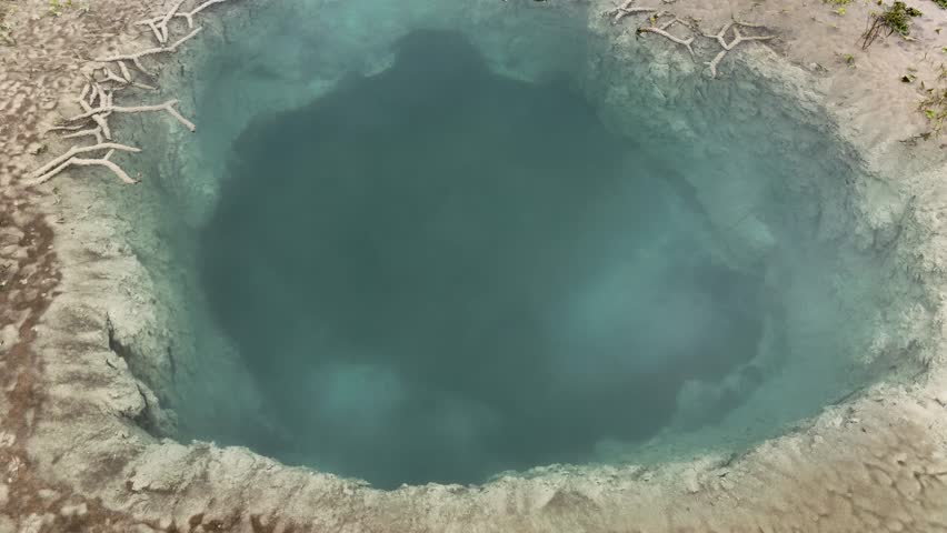 High angle view of a circular thermal water pond with deep turquoise colors surrounded by mineral deposits and dry branches creating a unique geothermal eye shape
