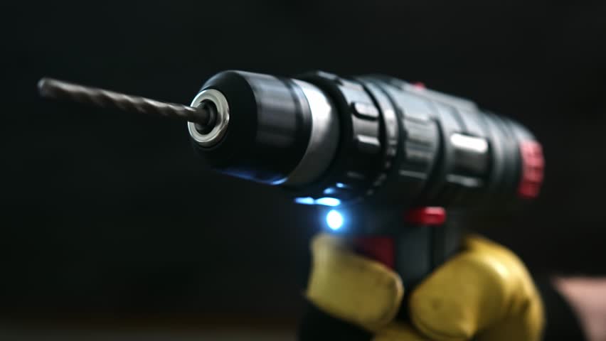 Hand grips a cordless drill with a metal bit in a workshop setting. Bright light shines from the tool.