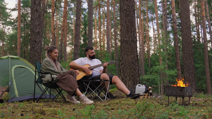 Young romantic couple enjoying a camping trip in the woods, with the man playing an acoustic guitar for his girlfriend while sitting near a cozy campfire and a tent in a pine forest