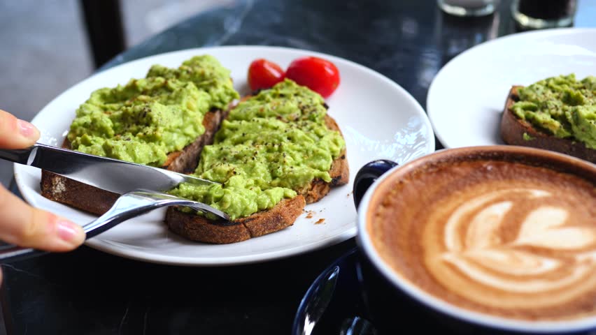Close up of a person cutting into a slice of avocado toast with a knife and fork, enjoying a healthy and trendy brunch with a cup of cappuccino at a cafe table outdoors