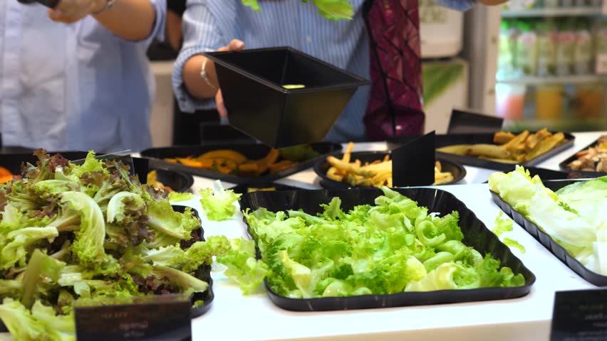 Customer using tongs to pick up fresh green lettuce and various vegetables from a self service salad bar counter in a grocery store, filling a bowl for a healthy takeaway meal