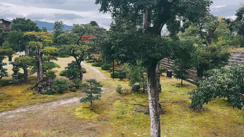 A carefully maintained japanese garden features sculpted pine trees, moss-covered ground, and gently curving gravel paths weaving through layered greenery. Stone elements and pruned tree forms create balance and intention, while distant mountains and soft clouds frame the scene, adding depth, calm, and a sense of timeless harmony.