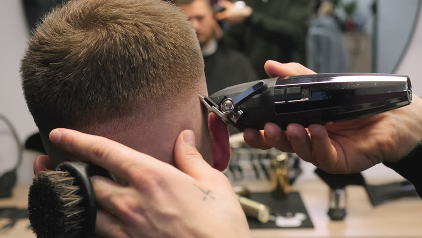 Hairdresser cuts man hair over ear with wireless trimmer in barbershop closeup. Professional barber serves male client doing haircut