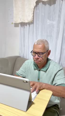 Elderly man with glasses concentrating while typing on a tablet keyboard, working from home