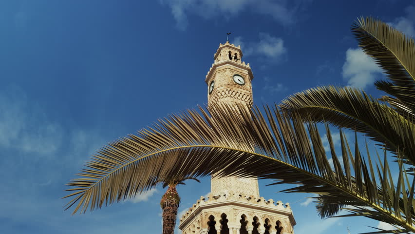 The iconic Izmir Clock Tower is seen through palm leaves in the city of Izmir, Turkiye, creating a layered urban scene that blends nature, history, and Mediterranean atmosphere.
