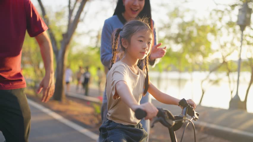 Asian young girl riding bicycle in garden while her parents look after. Adorable child feel happy and relax while learn to ride with family support, enjoying outdoor exercise activity in a public park