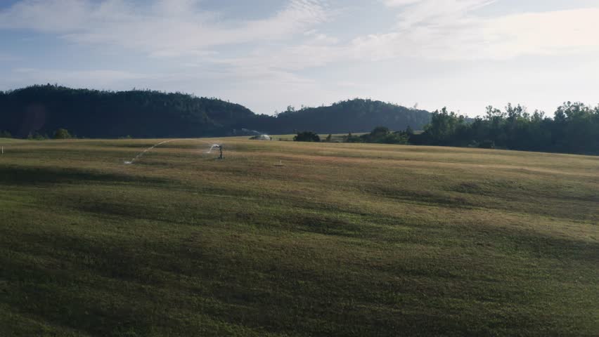 Aerial view of a manicured golf course with rolling green fairways, irrigation sprinklers, and forested hills.
