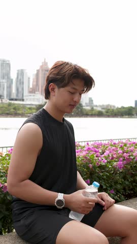 A fit man in sportswear drinks from a plastic bottle while sitting outdoors in daylight