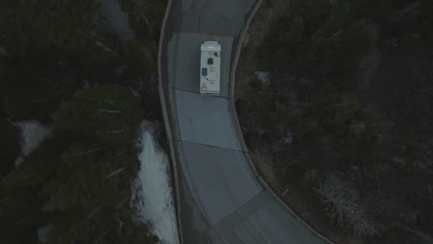 Top down aerial follow shot of a recreational vehicle driving along a winding mountain road through forest. Cinematic drone footage showing a road trip journey and exploration in a natural wilderness setting.
