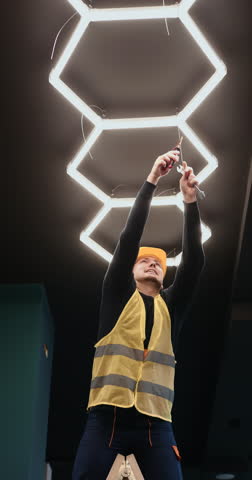 Electrician installing modern hexagon LED ceiling lighting in contemporary interior, using tools and tablet during electrical setup. Professional indoor renovation and smart lighting installation.