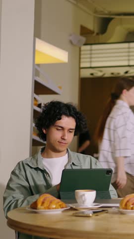 Young man with curly hair using digital tablet while sitting alone at round cafe table with coffee and croissants in cozy modern coffee shop interior
