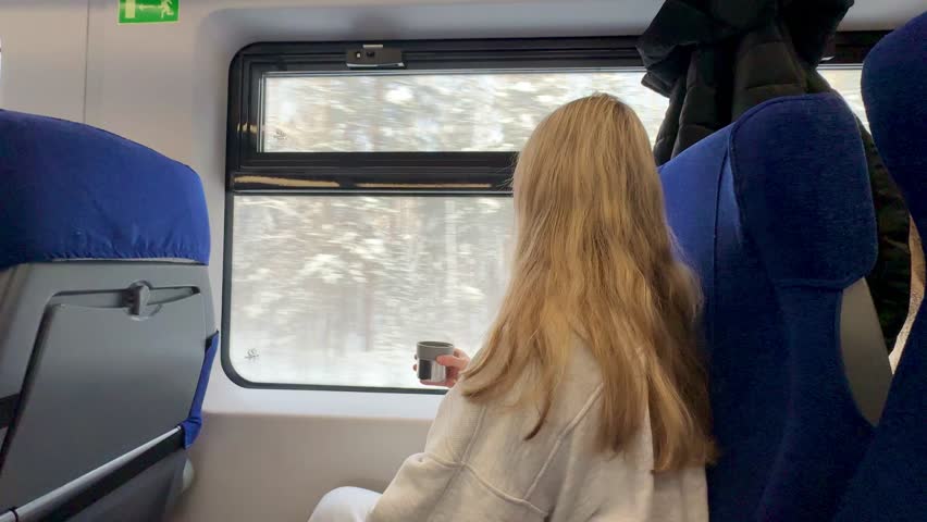 A young, happy blonde girl is traveling in winter, sitting on a train, looking out the window and drinking hot tea.Video 4k 