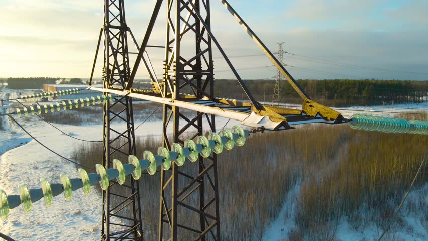 Drone flies beside icy power line insulators on a snowy transmission tower above frozen fields.