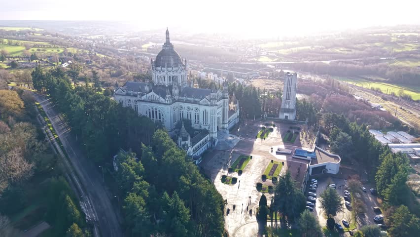 Drone pulling out from Basilique Sainte-Thérèse de Lisieux in Lisieux, Calvados, Normandy, France, revealing the Esplanade Sainte-Thérèse, Jardin du Carmel park and surrounding city and countryside