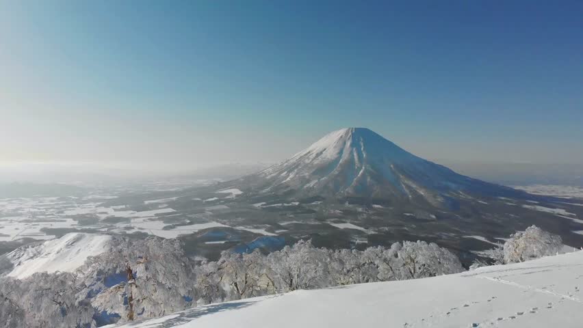 Scenic view from snowy mountaintop of Mount Yotei during bright winter day. People are hiking and enjoying landscape. Snow-covered trees and blue sky complete picture.