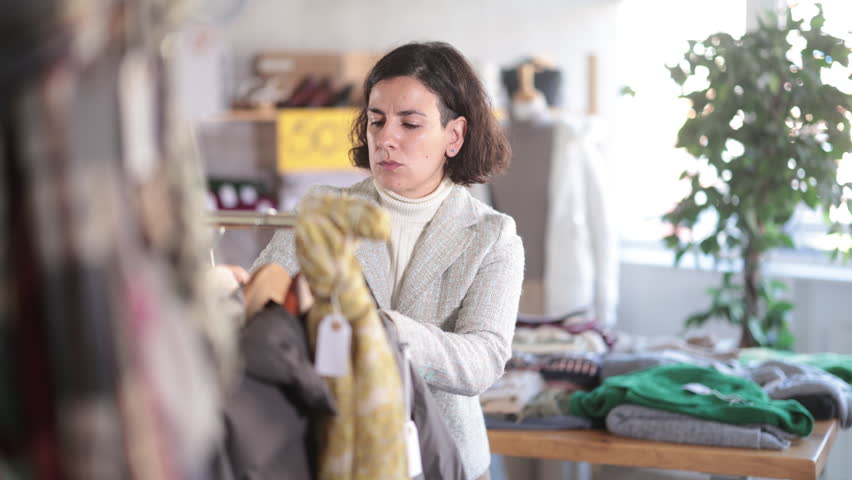 Adult woman stands near the rail with outerwear and chooses jackets and coats for the autumn and winter period. Buyer at a bargain sale of warm clothes. High quality 4k footage