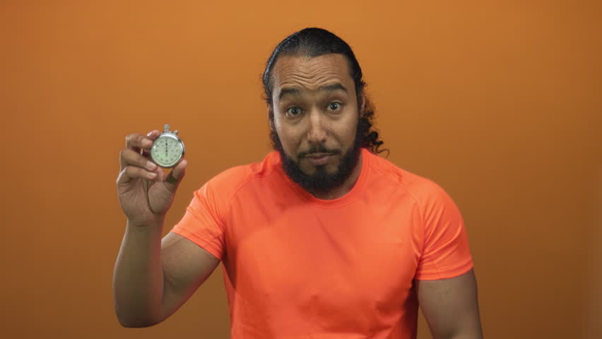 Man holding stopwatch and pointing with bare hand in orange studio, smiling and showing timer to camera; urgency time management motivation.
