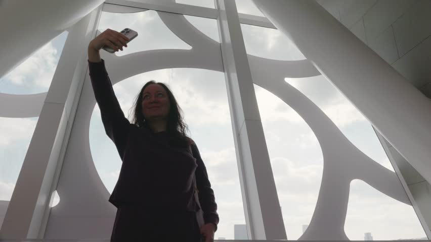 Middle aged caucasian woman standing indoors and taking selfie with smartphone at modern observation deck, city skyline visible through panoramic window, symbolizing tourism personal travel memories.