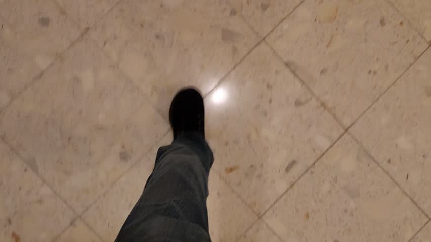 Video shot downward shows male legs wearing blue jeans and black sneakers moving from tiled floor onto carpet and walking quickly along a corridor The scene represents indoor movement transition between floor surfaces