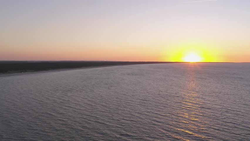 Picturesque sunset over the Baltic Sea. The low sun is reflected in the calm sea, while the endless sandy beach and the adjacent coastal forest are bathed in warm, golden light.