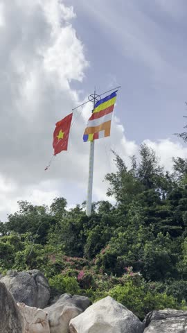 Two flags wave on a tall pole above a green hill with rocks in the foreground. The red flag with a yellow star and the colorful Buddhist flag create a vibrant scene against a partly cloudy sky.